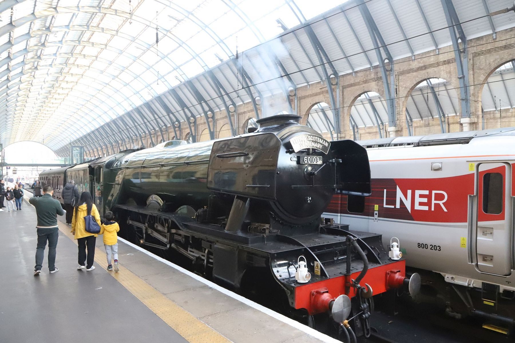 Family looking at Flying Scotsman next to an Azuma. Flying Scotsman steam locomotive at King's Cross railway station platform 8 for the 170th anniversary of the station's opening and the start of Flying Scotsman's 100th birthday celebrations